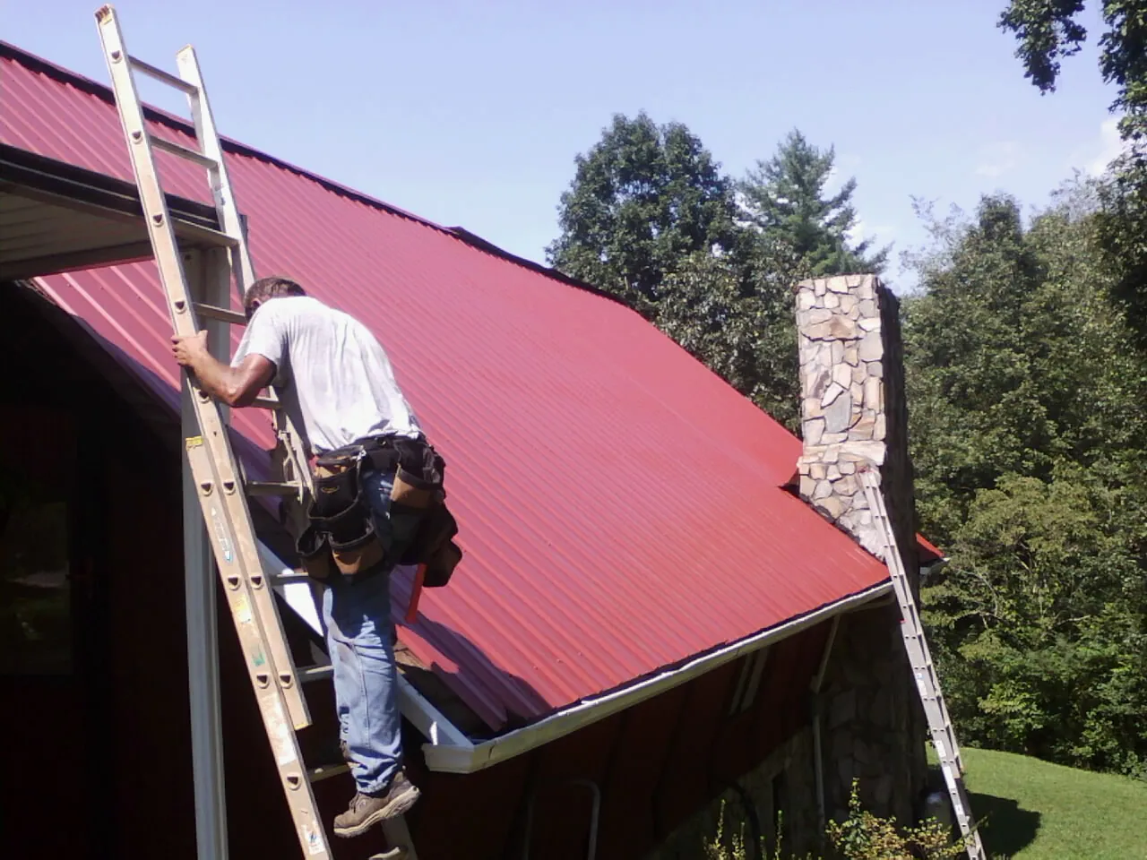 Roofer on a ladder beside a red metal roof and stone chimney — Soffit & Fascia Repair by Roofing Brothers in Russell